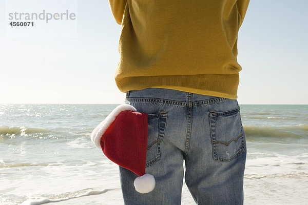 Mann am Strand stehend mit Blick auf den Strand  Nikolausmütze in der Gesäßtasche