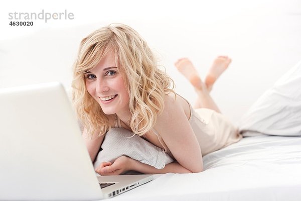 Junge Frau auf dem Bett liegend und mit Laptop  lächelnd  Porträt