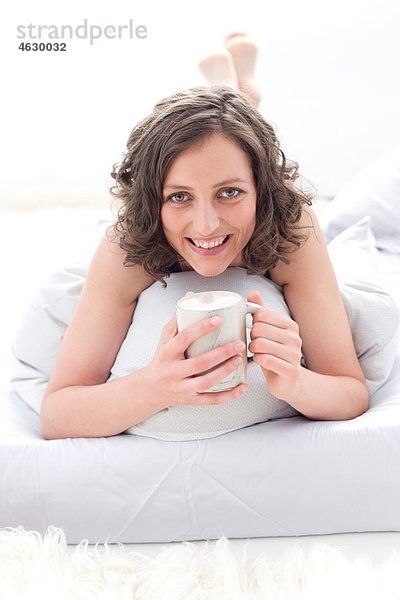 Junge Frau auf dem Bett liegend mit Kaffeetasse  lächelnd  Porträt