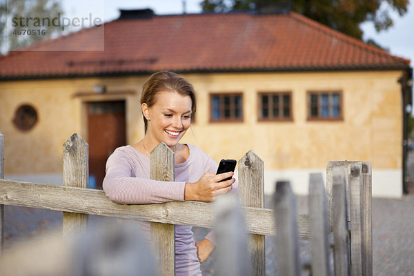 Glückliches Mädchen  das sich mit dem Handy in der Hand gegen den Zaun lehnt.