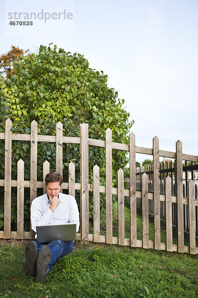 Mann  der sich gegen den Zaun lehnt  mit einem Computer auf dem Schoß.