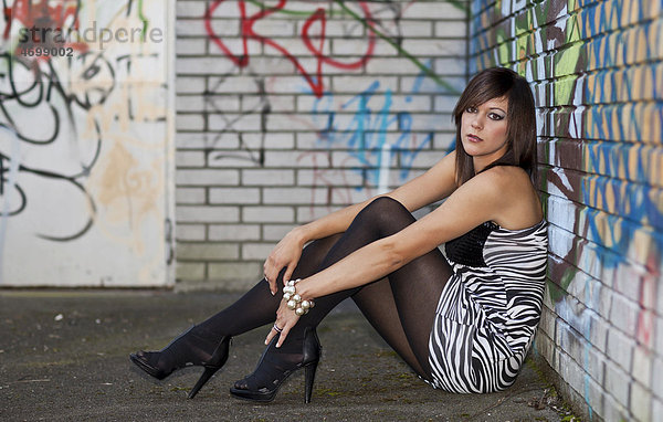 Junge Frau mit dunklen Haaren  Oberteil mit Zebra-Muster und hochhackigen Schuhen posiert sitzend vor Graffitiwand