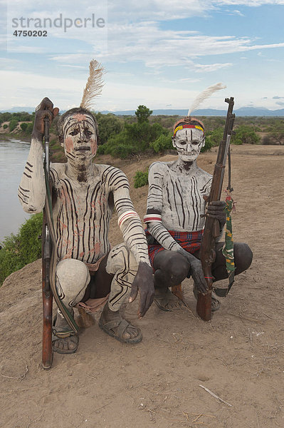 Zwei Karo Krieger mit Körperbemalung und Gesichtsbemalung und Gewehr sitzen auf ihrer Kopfstütze  Omo-Tal  Südäthiopien  Äthiopien  Afrika