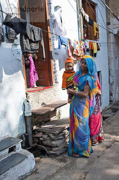 Junge freundliche indische Frau im Sari steht mit einem kleinen Kind auf ihrem Arm vor einem Hauseingang  Udaipur  Rajasthan  Indien  Asien