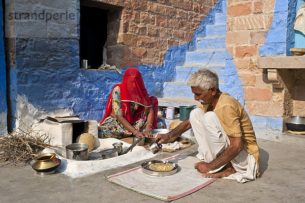Inderin mit Schleier backt an einem kleinen Ofen im Innenhof ihres Hauses Fladenbrot  Chapati  ihr Mann gießt darüber Ghee  zerlaufene Butter  Volksgruppe der Bishnoi  Jodhpur  Rajasthan  Indien  Asien