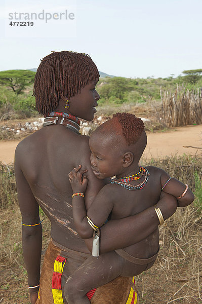 Junge Hamar Frau mit traditioneller Haarfrisur aus rotem Lehm mit Baby im Arm und mit Narben von Stockhieben auf dem Rücken  Omo-Tal  Süd Äthiopien  Afrika