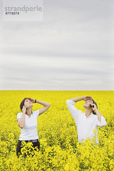 Junges Paar steht in einem Rapsfeld  telefoniert und schaut dabei zum Himmel