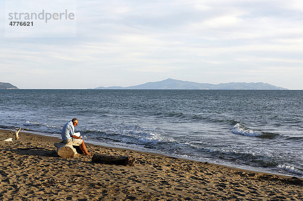 Alter Mann sitzt auf Treibholz am Mittelmeerstrand und liest  Maremma  Insel Giglio in der Ferne  Toskana  Italien  Europa