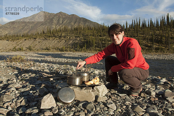 Junge Frau kocht auf einem Lagerfeuer  Kiesbank  dahinter die Northern Mackenzie Mountains und der Wind River  Yukon Territory  Kanada