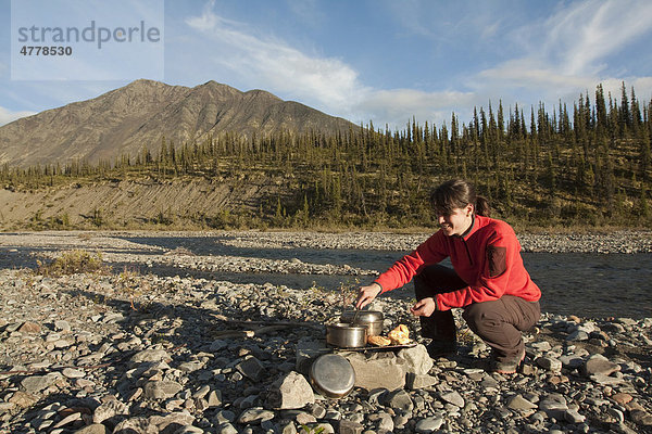 Junge Frau kocht auf einem Lagerfeuer  Kiesbank  dahinter die Northern Mackenzie Mountains und der Wind River  Yukon Territory  Kanada