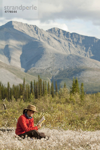 Junge Frau sitzt entspannt im Gras  Wollgras  und liest ein Buch  Northern Mackenzie Mountains  Wind River  Yukon Territory  Kanada