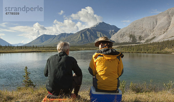 Paar  Mann und Frau sitzen am Ufer und genießen den Blick über den Fluss  Wind River Valley  nördliche Mackenzie Mountains Gebirgskette  Yukon Territorium  Kanada
