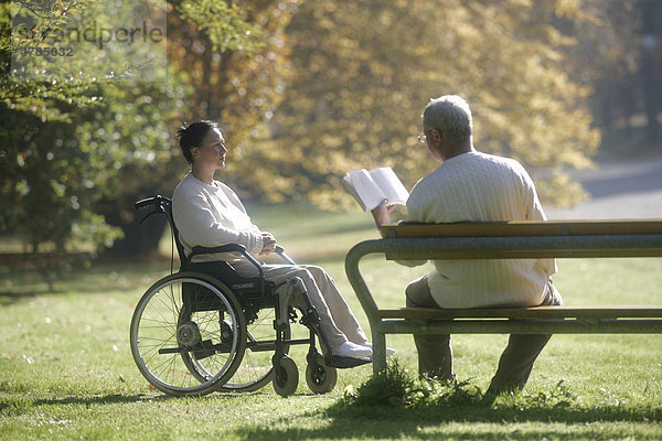 Frau sitzt im Rollstuhl  Mann sitzt mit Buch auf Parkbank