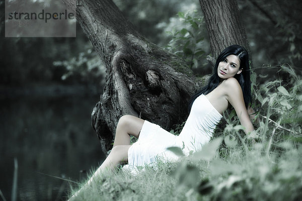 Junge Frau in einem weißen Kleid sitzt umrahmt von Bäumen an einem See
