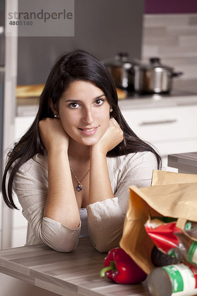 Junge Frau sitzt in der Küche am Esstisch  im Vordergrund eine Papiertüte mit Einkäufen