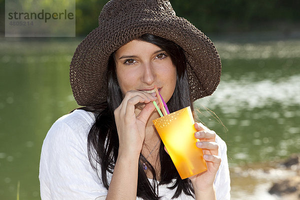 Junge Frau mit Strohhut sitzt am Strand eines Sees und trinkt ein Erfrischungsgetränk