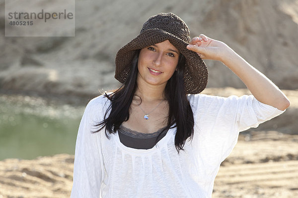 Junge Frau mit Strohhut am Strand eines Baggersees