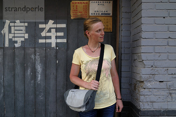 Frau mit roten Haaren und Umhängetasche steht vor einem Haus im chinesischen Hutong mit chinesischen Schriftzeichen  Peking  China  Asien