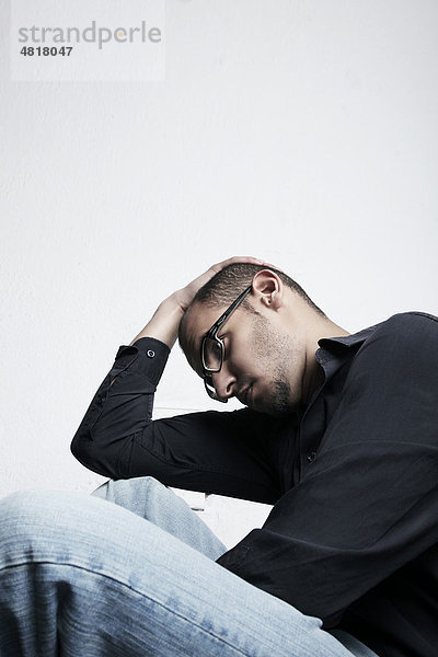 Junger Mann mit Brille sitzt auf dem Boden und hält sich die Hand an den Kopf