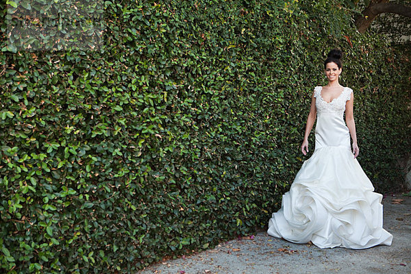 Junge Frau im Brautkleid  an der Hecke stehend