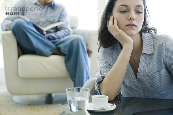 Frau stützt sich auf den Ellenbogen und sieht traurig aus  Mann liest auf dem Sofa im Hintergrund.