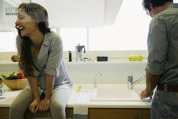Paar in der Küche  Mann beim Geschirrspülen  Frau auf der Theke sitzend  lachend
