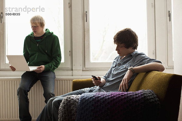 Ein Teenager auf einem Laptop  während sein Freund SMS-Nachrichten auf seinem Handy sendet.