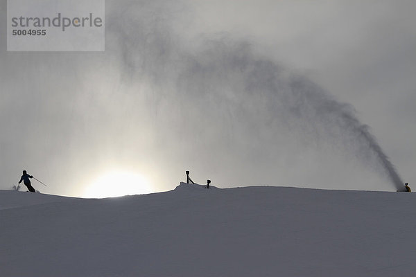 Eine Schneemaschine  die den Schnee von einem Skifahrer auf eine Piste spritzt.