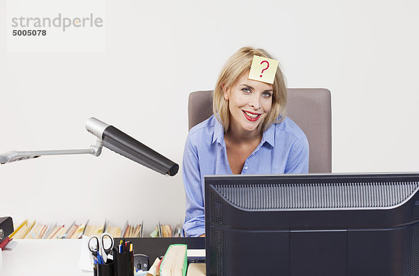 Eine Frau sitzt an einem Schreibtisch mit einem Klebezettel auf der Stirn.