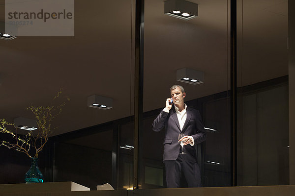 Gut gekleideter Mann schaut durchs Fenster  während er am Telefon ein Glas Wein hält.