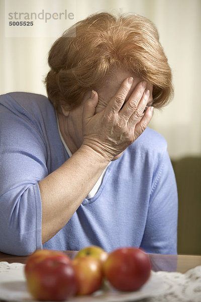 Eine ältere Frau sitzt an einem Tisch und bedeckt ihr Gesicht mit ihrer Hand.
