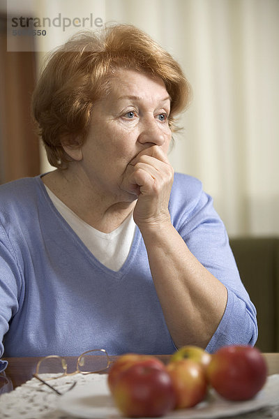 Eine besorgte ältere Frau  die mit der Hand am Tisch sitzt und ihren Mund bedeckt.