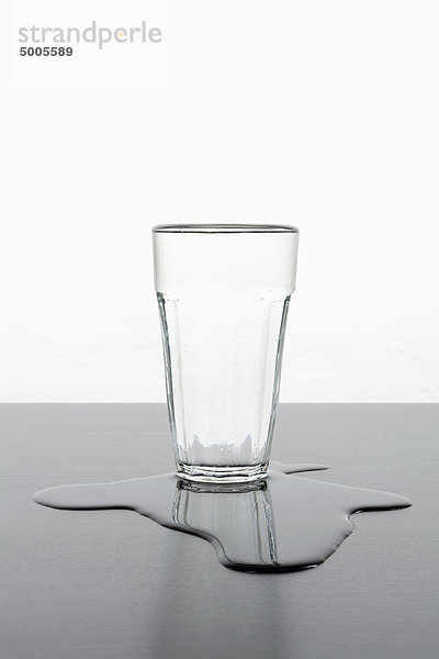 Ein aufrechtes Glas  das in einer Pfütze mit verschüttetem Wasser steht.