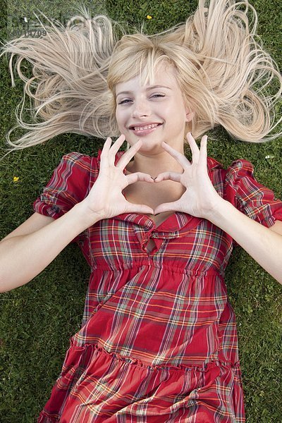 Lächelnde blonde Frau im Gras formt ein Herz mit ihren Händen