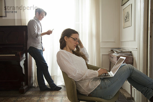 Frau mit Laptop zu Hause  Mann mit Handy im Hintergrund