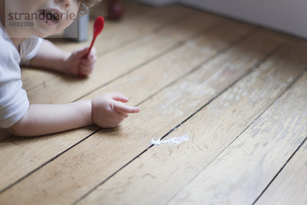 Kleinkind auf dem Boden liegend mit Löffel in der Hand  auf verschüttetes Essen zeigend
