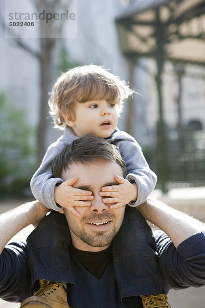 Vater trägt Kleinkindesohn auf seinen Schultern  Sohn bedeckt Vaters Augen mit seinen Händen.