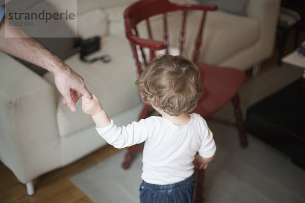 Kleinkind hält sich an Vaters Hand fest  geht auf das Sofa zu  abgeschnitten