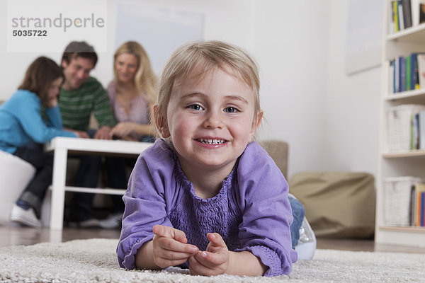 Mädchen auf Teppich liegend mit Familie im Hintergrund sitzend