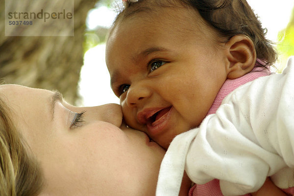 Caucasian Mutter halten Sie die up lächelnd mixed Race weiblichen Baby und küssen ihr Gesicht