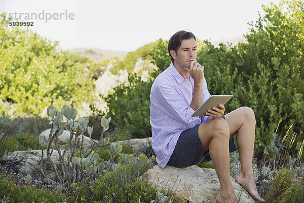 Mann sitzt auf einem Felsen mit einem digitalen Tablett und denkt nach.