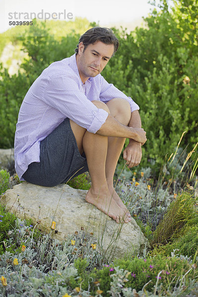 Ein erwachsener Mann  der auf einem Felsen in einem Garten sitzt und nachdenkt.