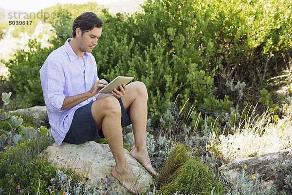 Mittlerer Erwachsener Mann  der auf einem Felsen sitzt und ein digitales Tablett benutzt.