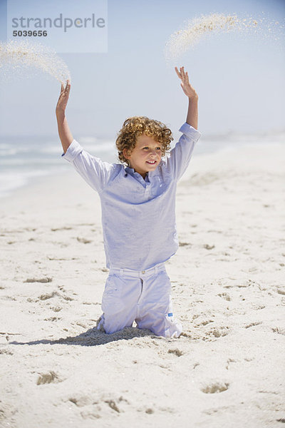 Junge  der mit erhobenen Armen im Sand spielt.