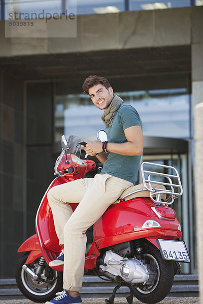 Portrait eines jungen Mannes  der auf einem Roller sitzt und Textnachrichten verschickt.