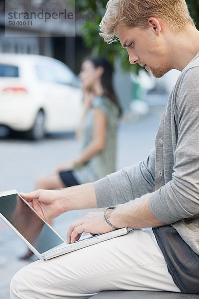 Junger Mann mit einem Laptop und einer Frau im Hintergrund auf einer Straße
