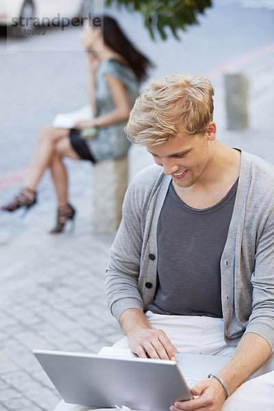 Junger Mann mit einem Laptop und einer Frau im Hintergrund auf einer Straße