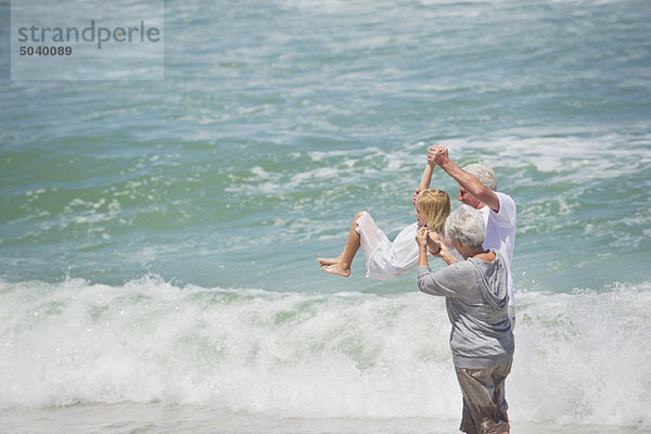 Seniorenpaar hebt ihre Enkelin am Strand in die Luft