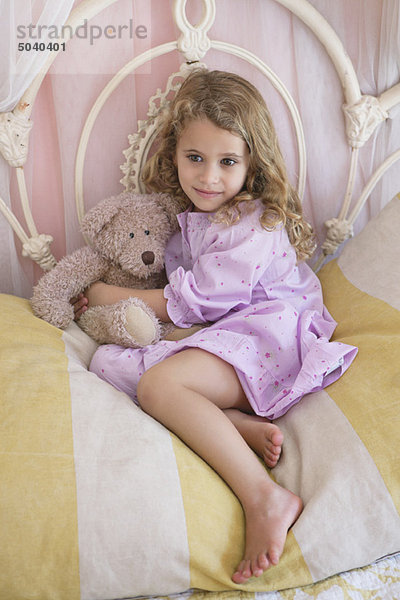 Süßes kleines Mädchen mit Teddybär auf dem Bett sitzend