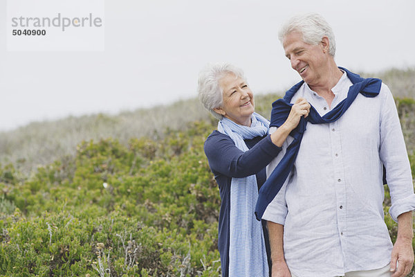 Ältere Frau  die am Strand einen Pullover auf die Schulter ihres Mannes legt.
